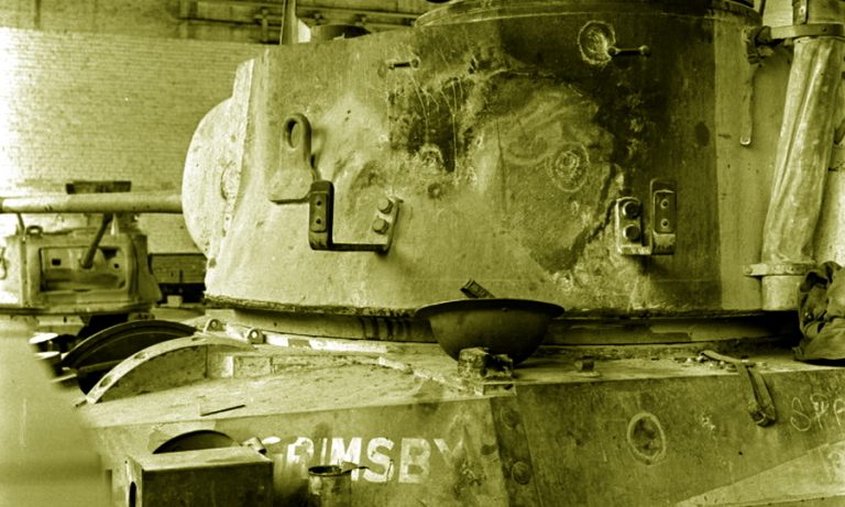 Следы попаданий в башенную и бортовую броню танка с личным именем «Гримсби» хорошо видны в виде круглых отпечатков. Снимок сделан в ремонтных мастерских