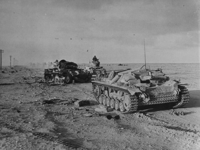 Подбитые танк Pz.Kpfw.III и штурмовое орудие StuG III Ausf.D (справа) из состава SonderVerband 288 на дороге в Северной Африке. Мимо проезжает колонна британских танков M3 «Stuart» Источник – waralbum.ru