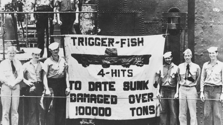  Члены экипажа SS-237 «Триггер» на фоне закреплённого на рубке подлодки плаката, сообщающего о четырёх торпедных попаданиях во вражеский авианосец.